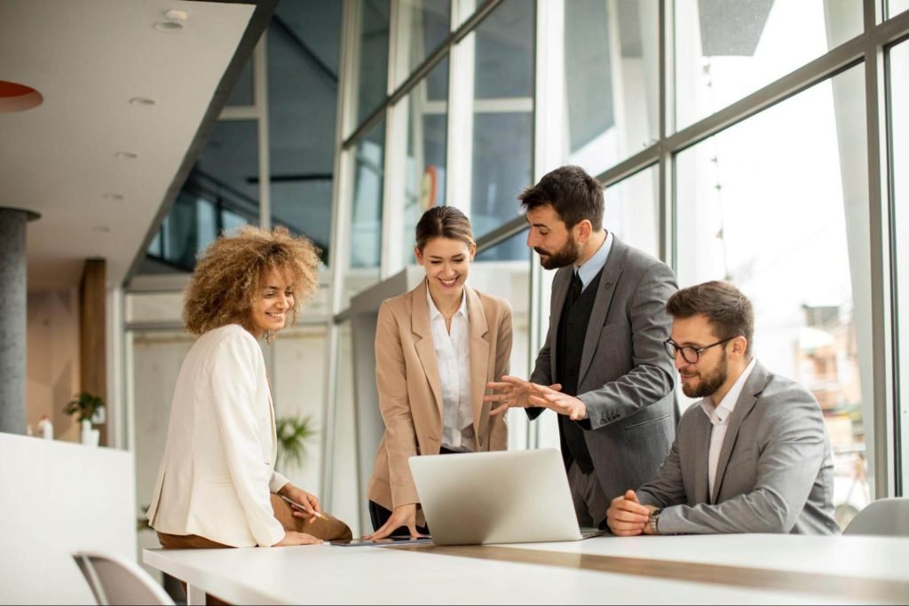 A diverse group of four professionals engaging in a discussion while looking at a laptop in a modern office space, showcasing teamwork and collaboration.