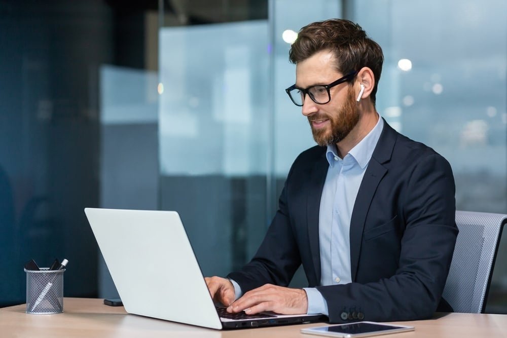 Businessman in formal attire using a laptop and wearing wireless earbuds during a video call in a modern office